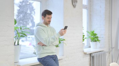 Young Adult Man using Smartphone while Standing Near Window