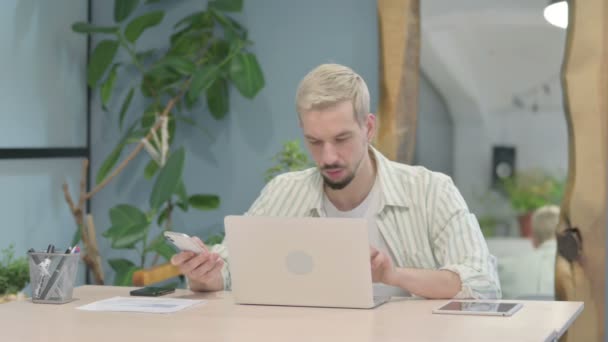 Σύγχρονος Νεαρός Άνδρας Που Χρησιμοποιεί Smartphone Ενώ Χρησιμοποιεί Laptop — Αρχείο Βίντεο