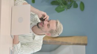 Genç adamın telefonda konuştuğu ve dizüstü bilgisayar kullandığı dikey video.
