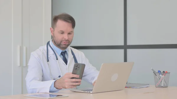 在笔记本电脑上工作并使用智能手机的年轻医生 — 图库照片