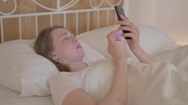 ベッドで寝そべっている間にスマートフォンでショックを受けた若い女性 — ストック動画
