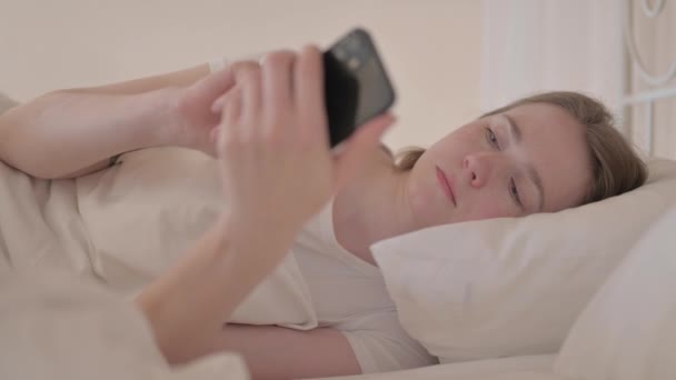 ベッドでエルボーに座っている間にスマートフォンを使用して若い女性のサイドビュー — ストック動画