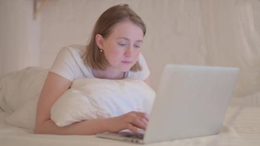 Dizüstü bilgisayarda kutlama yapan genç bir kadın yatakta karın üstü yatıyor.