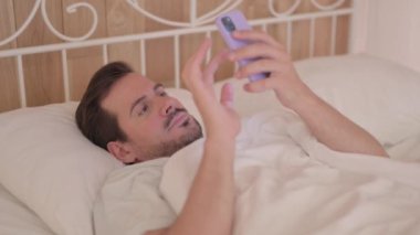 Genç adam yatakta yatarken telefonu kullanıyor.