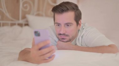Genç adam karnının üstünde yatarken akıllı telefon kullanıyor.