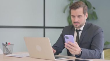 Dizüstü bilgisayarda çalışan ve akıllı telefon kullanan genç bir iş adamı.