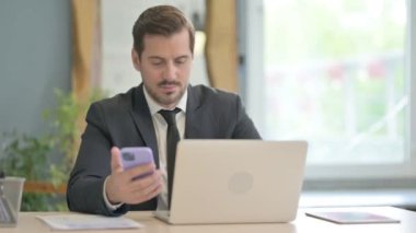 Dizüstü bilgisayarda çalışan ve akıllı telefon kullanan genç bir iş adamı.