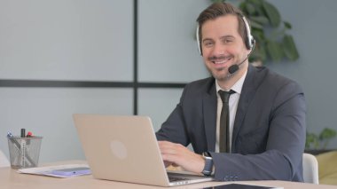 Çağrı merkezinde kameraya gülümseyen kulaklıklı bir iş adamı.