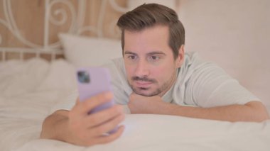 Genç adam karnının üstünde yatarken akıllı telefon kullanıyor.