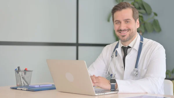 ノートパソコンで働く男性医師がカメラで微笑む — ストック写真