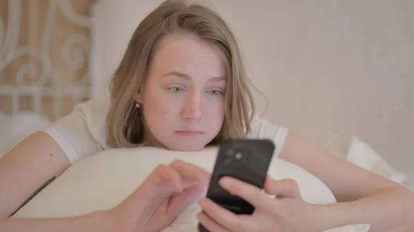 Młoda Kobieta Korzystająca Smartfona Podczas Leżenia Brzuchu Łóżku — Zdjęcie stockowe