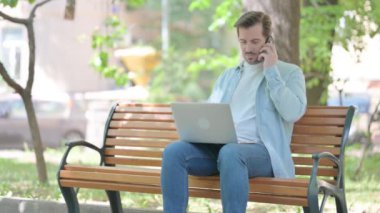 Kızgın genç adam dışarıda dizüstü bilgisayar kullanırken telefonla konuşuyor.