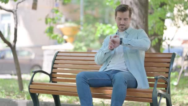 Junger Mann Benutzt Smartwatch Während Draußen Auf Bank Sitzt — Stockvideo