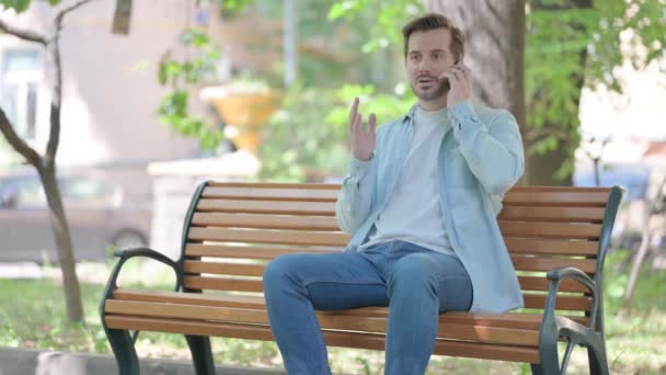 ベンチに屋外に座っている間 電話で主張する若い男 — ストック動画
