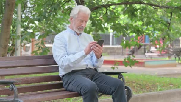 长者坐在户外的长椅上 一边用智能手机庆祝成功 — 图库视频影像