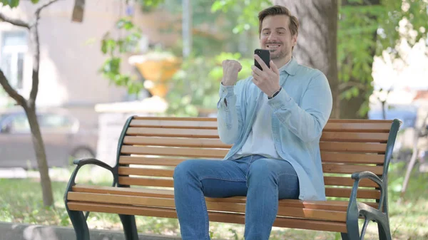 Jongeman Viert Succes Smartphone Tijdens Het Zitten Buiten Een Bank — Stockfoto