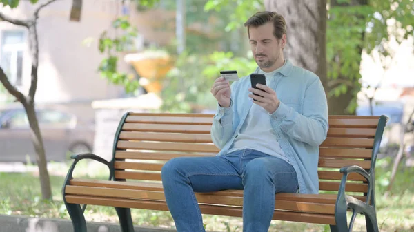 Młody Człowiek Robi Zakupy Online Telefon Podczas Siedzenia Zewnątrz Ławce — Zdjęcie stockowe