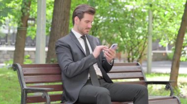 Bir bankta otururken Smartphone 'da Başarıyı Kutlayan Genç İşadamı