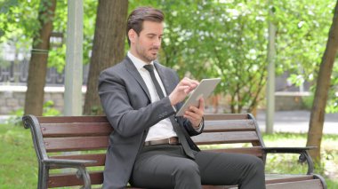 Tablet kullanan genç bir işadamı açık bir bankta otururken
