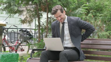 Orta yaşlı iş adamı dışarıda otururken dizüstü bilgisayarla sırt ağrısı çekiyor.