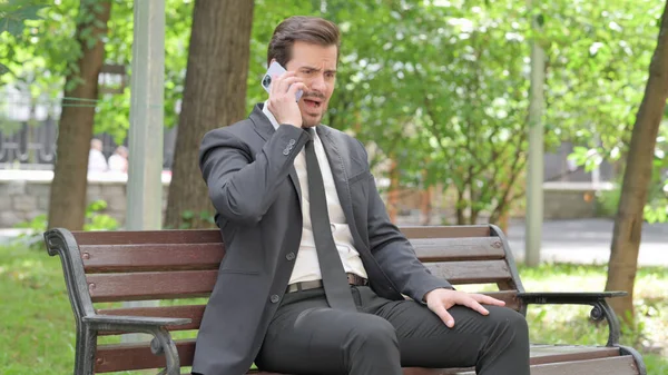 Zdenerwowany Młody Biznesmen Kłócący Się Przez Telefon Podczas Siedzenia Zewnątrz — Zdjęcie stockowe