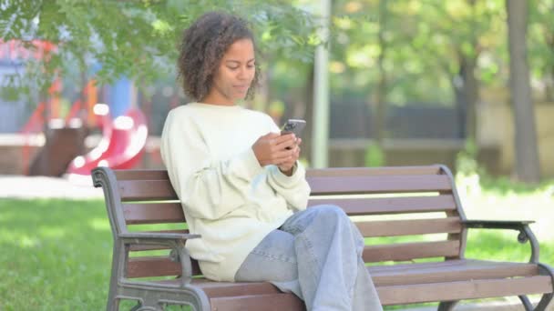 坐在室外长椅上的非洲年轻女子因电话失物招领而震惊 — 图库视频影像