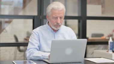 Ofiste dizüstü bilgisayarla çalışırken kameraya bakan eski iş adamı