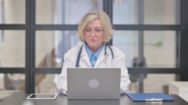 Baş parmakları aşağıda yaşlı kadın doktor kameraya bakıyor.