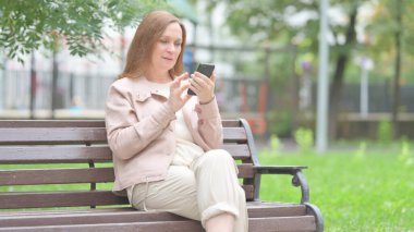 Yaşlı kadın bankta otururken telefonu kullanıyor.