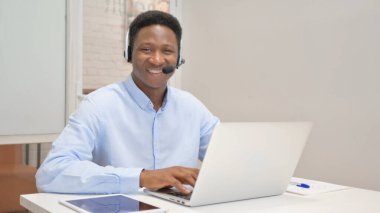 Kulaklıklı gülümseyen Afrikalı Çağrı Merkezindeki Kameraya Bakıyor