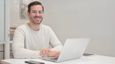 Ofiste dizüstü bilgisayarla çalışırken kameraya gülümseyen orta yaşlı adam.
