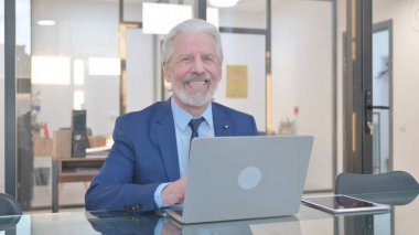 Ofisteki Dizüstü bilgisayarda çalışırken kameraya gülümseyen kıdemli iş adamı