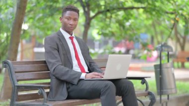 Şok olmuş Afrikalı iş adamı kucağında dizüstü bilgisayarla kameraya bakıyor.