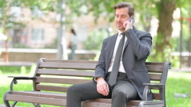 Orta yaşlı iş adamı dışarıda otururken telefonla konuşuyor.