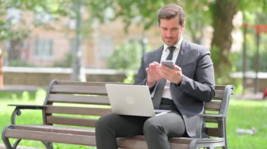 Orta yaşlı iş adamı telefonda konuşuyor ve dışarıda dizüstü bilgisayar kullanıyor.