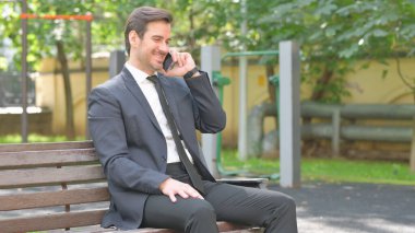 Genç İşadamı Bench 'te otururken telefonla konuşuyor.