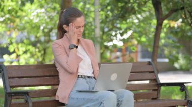 Migreni olan genç bir kadın parkta otururken dizüstü bilgisayar kullanıyor.