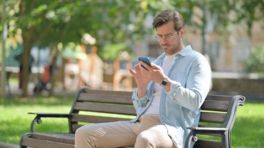 Sıradan genç adam parkta otururken akıllı telefon kullanıyor.
