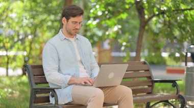 Sıradan genç adam parkta otururken dizüstü bilgisayar kullanıyor.