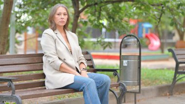 Yaşlı kadın bankta otururken kameraya bakıyor.