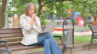 Dışarıda dizüstü bilgisayar kullanırken yaşlı hasta kadın öksürüyor.