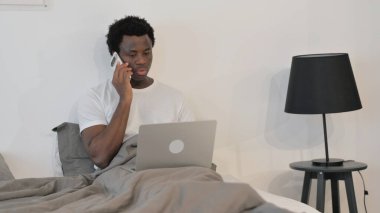 Afrikalı Adam telefonda dizüstü bilgisayarla yatakta konuşuyor.
