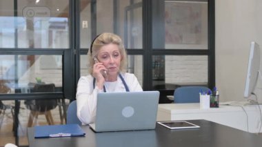 Klinikte dizüstü bilgisayar kullanırken telefonda konuşan yaşlı kadın doktor.