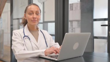 Bilgisayarda çalışırken kameraya gülümseyen çok ırklı kadın doktor.