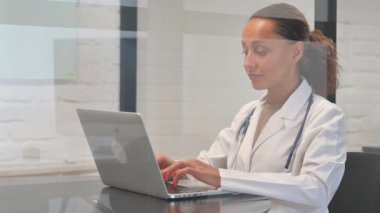 Bilgisayarda çalışırken kameraya gülümseyen çok ırklı kadın doktor.