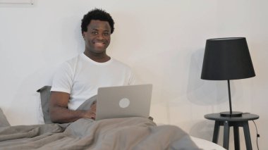 Yatakta dizüstü bilgisayarla kameraya bakan gülümseyen Afrikalı adam