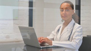 Bilgisayarda çalışırken kameraya bakan çok ırklı kadın doktor.