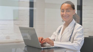Bilgisayarda çalışırken kameraya gülümseyen çok ırklı kadın doktor.