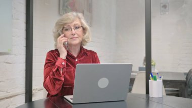 Dizüstü bilgisayarda çalışırken telefonda konuşan kıdemli yetişkin kadın.