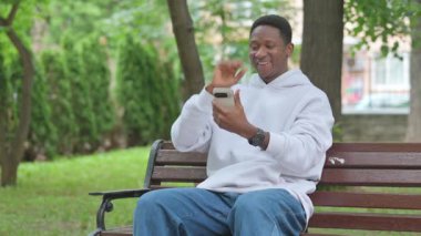 Genç Afrikalı bir adam telefonda görüntülü konuşma yapıyor, Bench 'in dışında oturuyor.